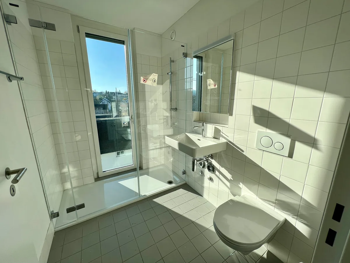 Man sieht von der Tür aus in ein Badezimmer. Rechts ist die Toilette, daneben das Waschbecken und im Hintergrund eine grosszügige Dusche, in die ein bodenlanges Fenster eingebaut ist.